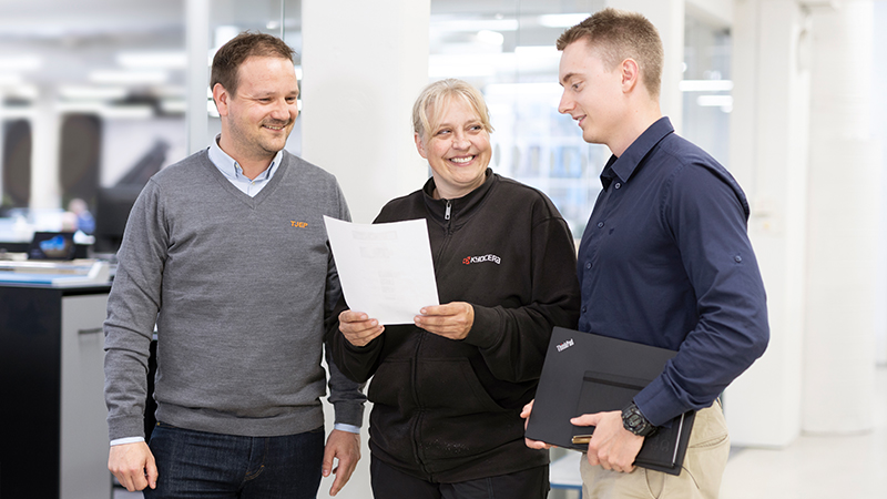 Three different colleagues standing in the middle of the office and the production area are having a dialogue on the basis of a piece of paper.