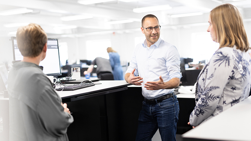 An employee is doing a presentation by his desk to his two colleagues