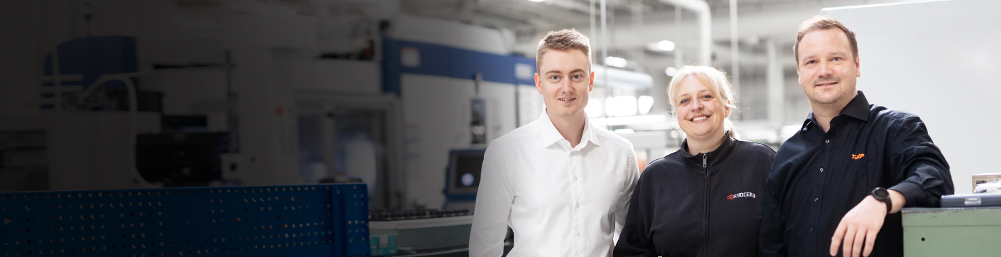 A group of three different employees are standing side by side in the production area, looking directly into the camera